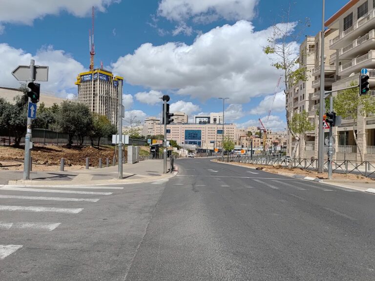 רחוב ירושלמי שומם מוקף בנייני מגורים ואתרי בנייה, על רקע שמיים כחולים עם עננים – סמל להאטה בשוק הנדל"ן בישראל בשנת 2025, על רקע דיונים גוברים בנושא ירידת מחירי הדירות בישראל 2025.