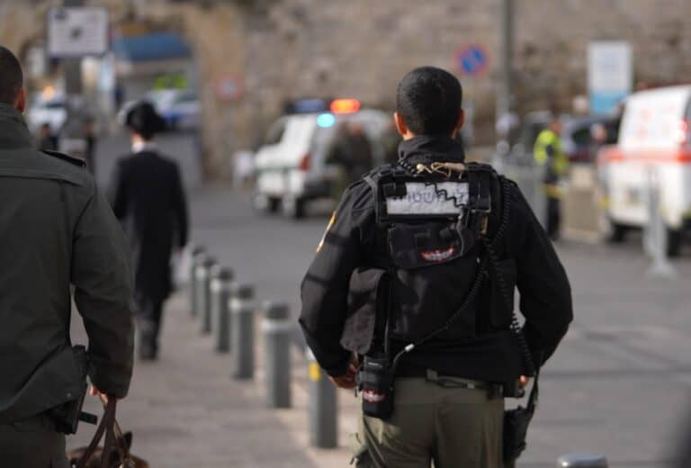 שוטרי מג"ב חמושים פוסעים ברחוב בעיר העתיקה בירושלים, ברקע רכבים ביטחוניים ותושבים – צילום המתאר את המתיחות הביטחונית במזרח העיר