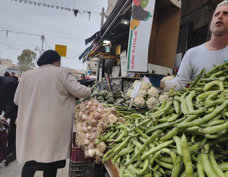 דוכן ירקות בשוק מחנה יהודה בירושלים עם פול ירוק, שום וירקות עונתיים נוספים, לקוחות וסוחר מקומי