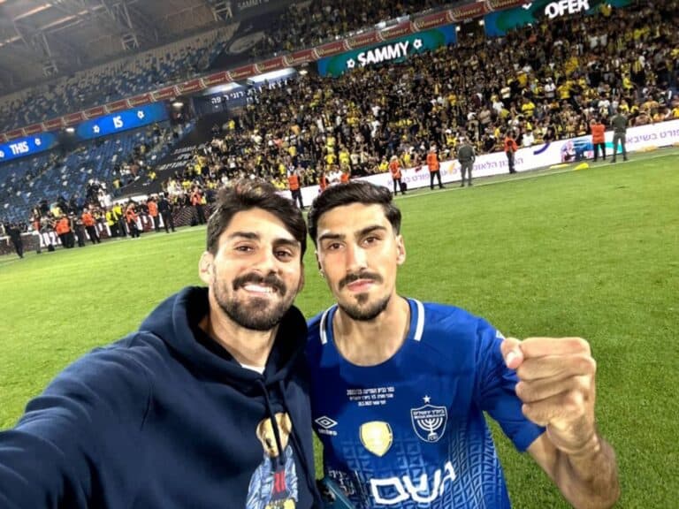 חזי וליאון מזרחי מצולמים יחד על כר הדשא, ברקע אוהדי בית"ר ביציע