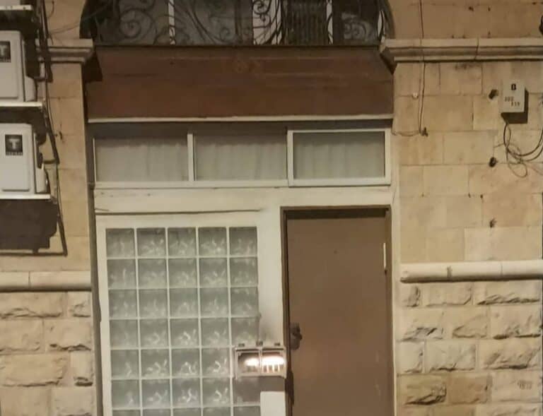 דלת ירושלמית סגורה, לצידה תיבת נרות מוארת – רמז לריח חמים שמתחיל בפנים