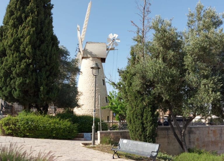 טחנת הרוח מונטיפיורי ההיסטורית במשכנות שאננים בירושלים, עם עצים וספסל ביום בהיר. סמל לבנייה היהודית מחוץ לחומות.