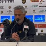 מאמן בית"ר ירושלים ברק יצחקי מדבר במסיבת עיתונאים עם לוגו הקבוצה ברקע.