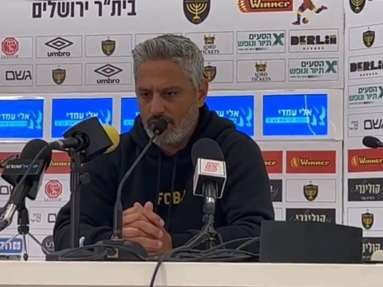 מאמן בית"ר ירושלים ברק יצחקי מדבר במסיבת עיתונאים עם לוגו הקבוצה ברקע.