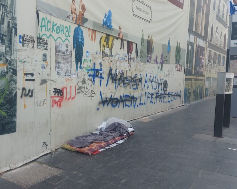 מראה של מקום לינה מאולתר (מזרן ושמיכות) על מדרכה בירושלים, המייצג את תופעת חסרי הבית והמצוקה ברחובות העיר.