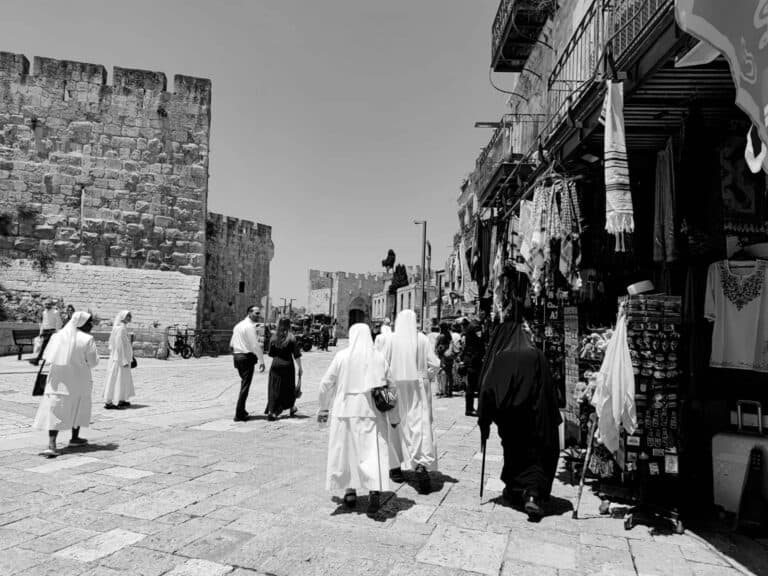 Black and white photo of a street in East Jerusalem with religious tourists and market stalls, reflecting the city’s diversity and vulnerability