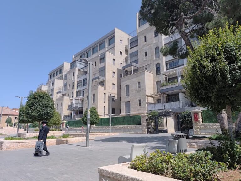 A modern, upscale residential complex in Jerusalem, possibly the former Schneller Camp, with a person appearing to be Haredi walking by