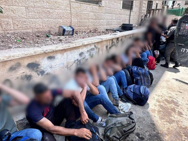 A group of men are sitting on the sidewalk with their hands on their heads, some are blurred, with a police van and a security force member standing nearby, likely after an arrest in Jerusalem